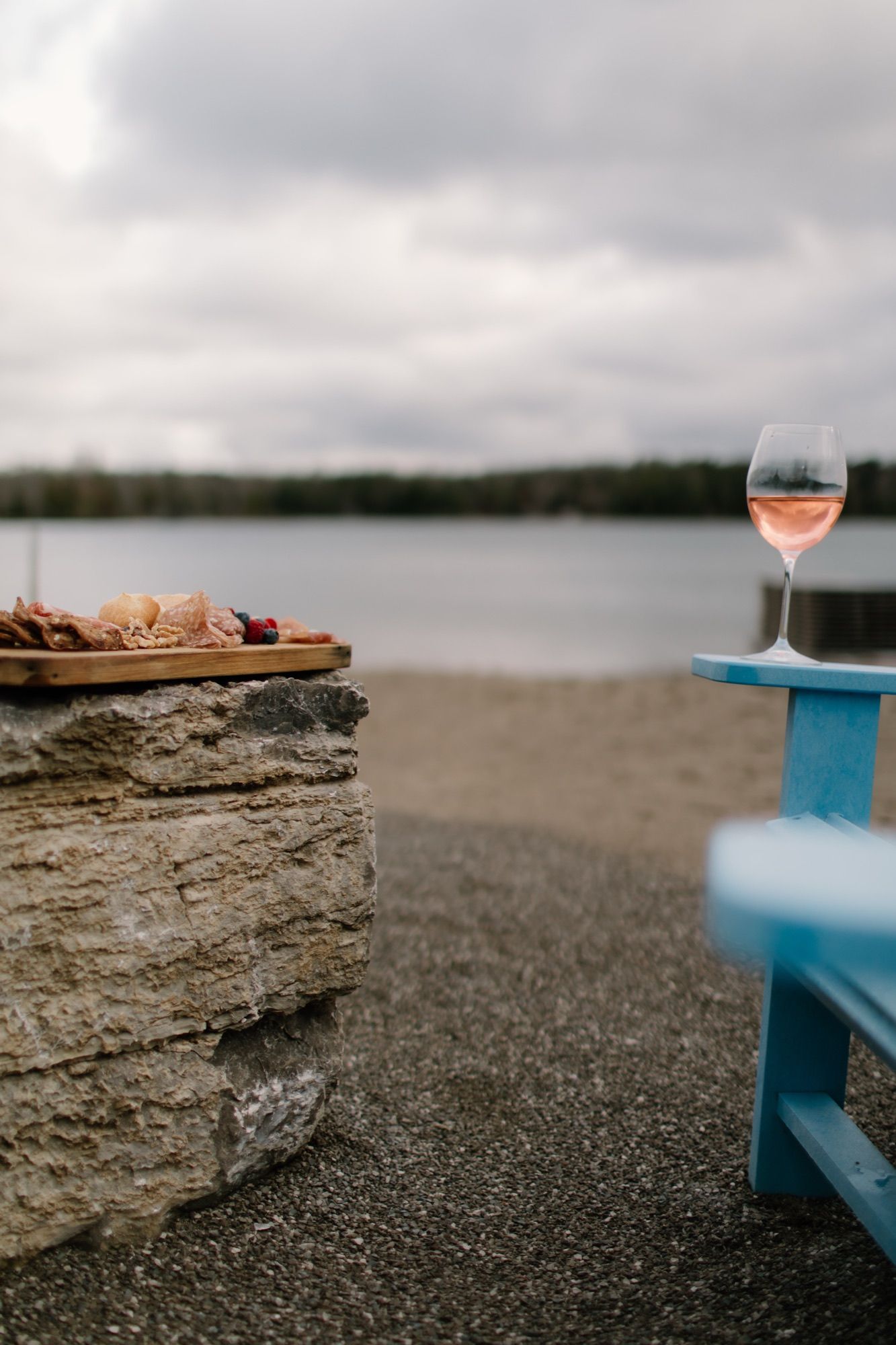 A charcuterie board on a stone ledge and a glass of rosé on the arm of a blue Adirondack chair, with the lake and its far shore behind.